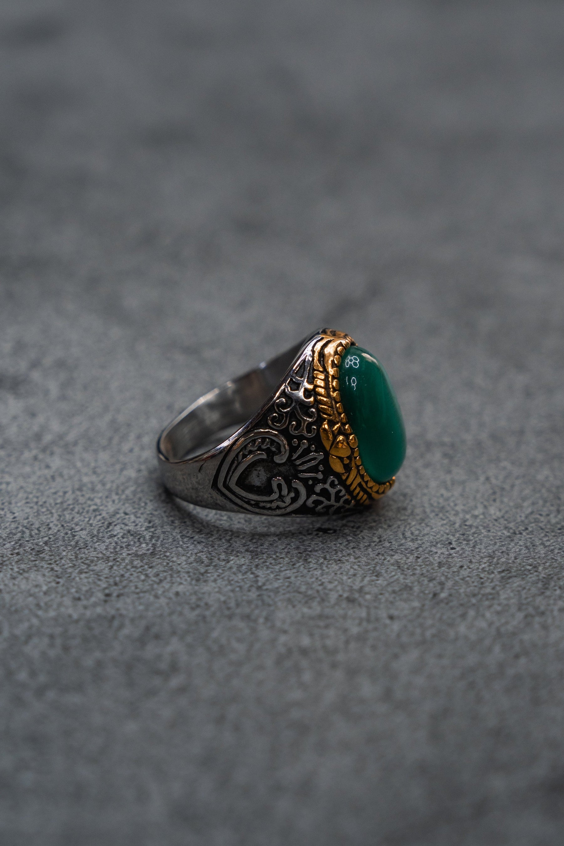 Displayed on a gray surface, The Relic Signet Ring by Relentless Betrayal features intricate engravings and gold accents, with a large deep green stone at its center; its weathered silver finish enhances the ring’s timeless elegance.