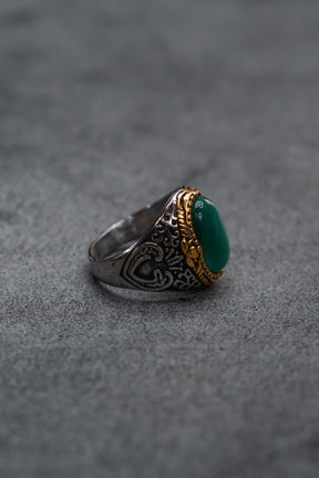 Displayed on a gray surface, The Relic Signet Ring by Relentless Betrayal features intricate engravings and gold accents, with a large deep green stone at its center; its weathered silver finish enhances the ring’s timeless elegance.