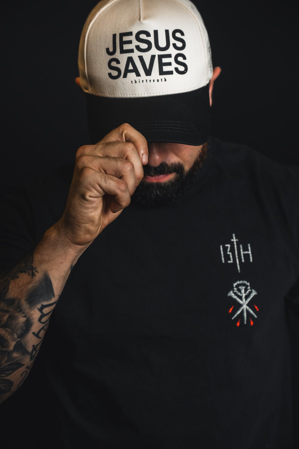 A man with tattoos, wearing a black shirt and the 13th Jesus Saves Ivory and Black Snapback, holds the brim of his embroidered baseball cap, partially covering his face.