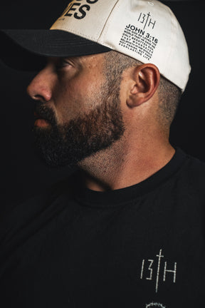 A bearded man wearing a black shirt and the 13th brand’s “Jesus Saves” Ivory and Black Snapback is shown in profile against a dark background.