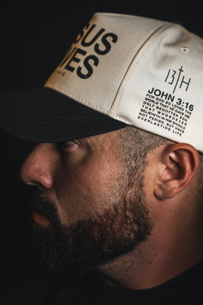 A bearded man in profile wears the 13th Jesus Saves Ivory and Black Snapback, a beige embroidered baseball cap featuring black text with the words JESUS LIVES and a John 3:16 Bible quote.