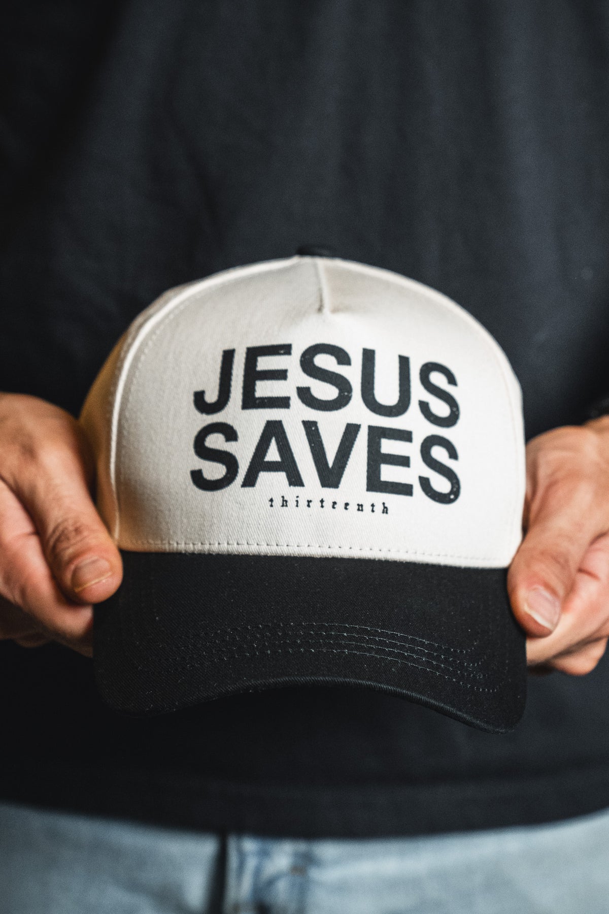 A person holds the Jesus Saves Ivory and Black Snapback by 13th, featuring “JESUS SAVES” and “13th” embroidered on the front.