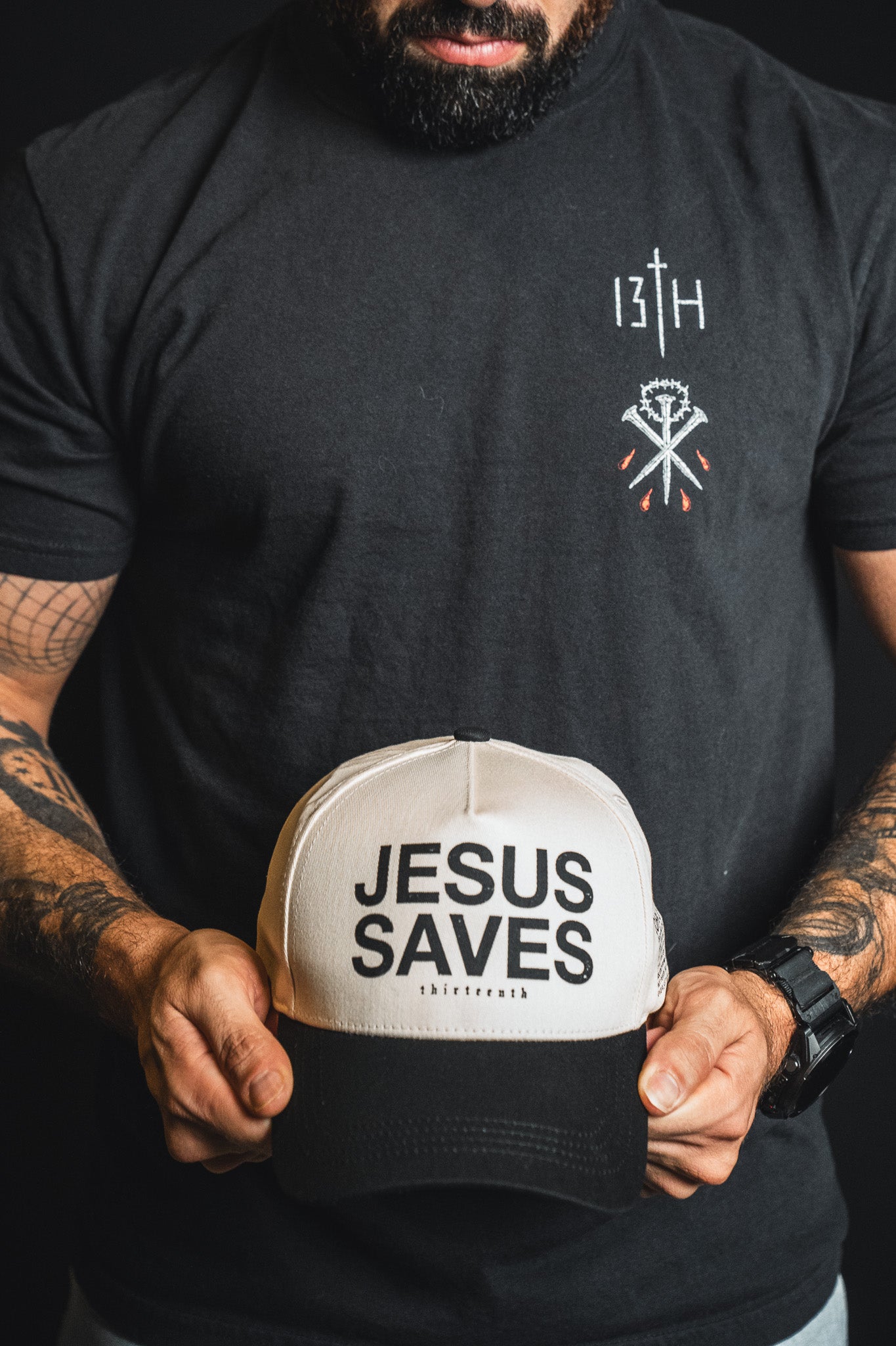 A person wearing a black t-shirt with small white and red chest designs holds the 13th Jesus Saves Ivory and Black Snapback, a bold-lettered hat from 13th.