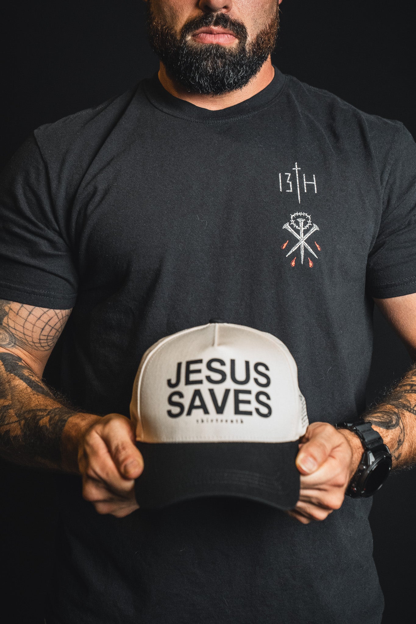 A bearded man holds the Jesus Saves Ivory and Black Snapback by 13th, wearing a black shirt with symbols and a watch, set against a dark background.