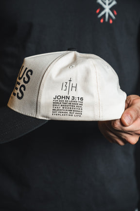 A person holds the Jesus Saves Ivory and Black Snapback by 13th, featuring white and black embroidery with “JOHN 3:16,” a Bible verse on the side, and bold “13th” branding—serving as a statement piece that reminds wearers Jesus saves.