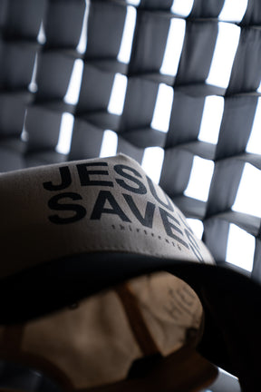 The 13th Jesus Saves Ivory and Black Snapback is displayed in front of a background featuring a geometric, grid-like light pattern.