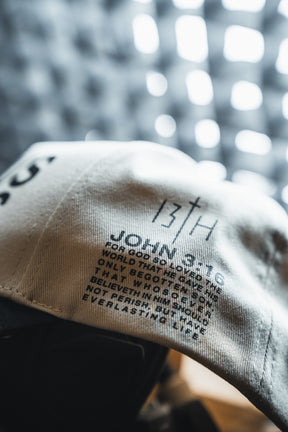 Close-up of a white fabric label with black printed text, featuring the 13th brand and motivational phrases, sewn onto the Jesus Saves Ivory and Black Snapback baseball cap.