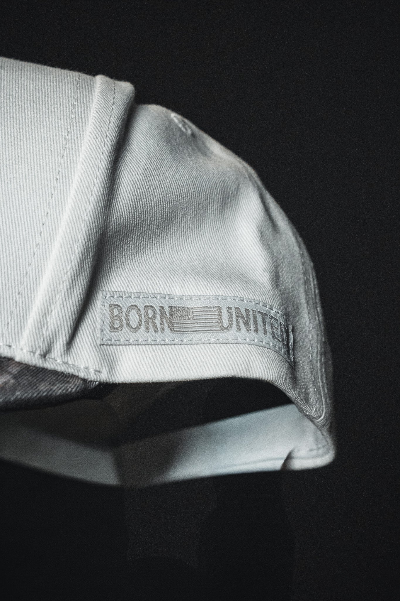 Close-up of the Born United Private Label “Jesus is King - Ivory and Deadwood Camo Snapback,” an embroidered baseball cap featuring the words BORN UNITED and a faded flag patch on the side, set against a dark background.