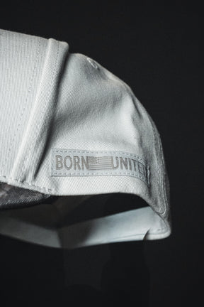 Close-up of the Born United Private Label “Jesus is King - Ivory and Deadwood Camo Snapback,” an embroidered baseball cap featuring the words BORN UNITED and a faded flag patch on the side, set against a dark background.