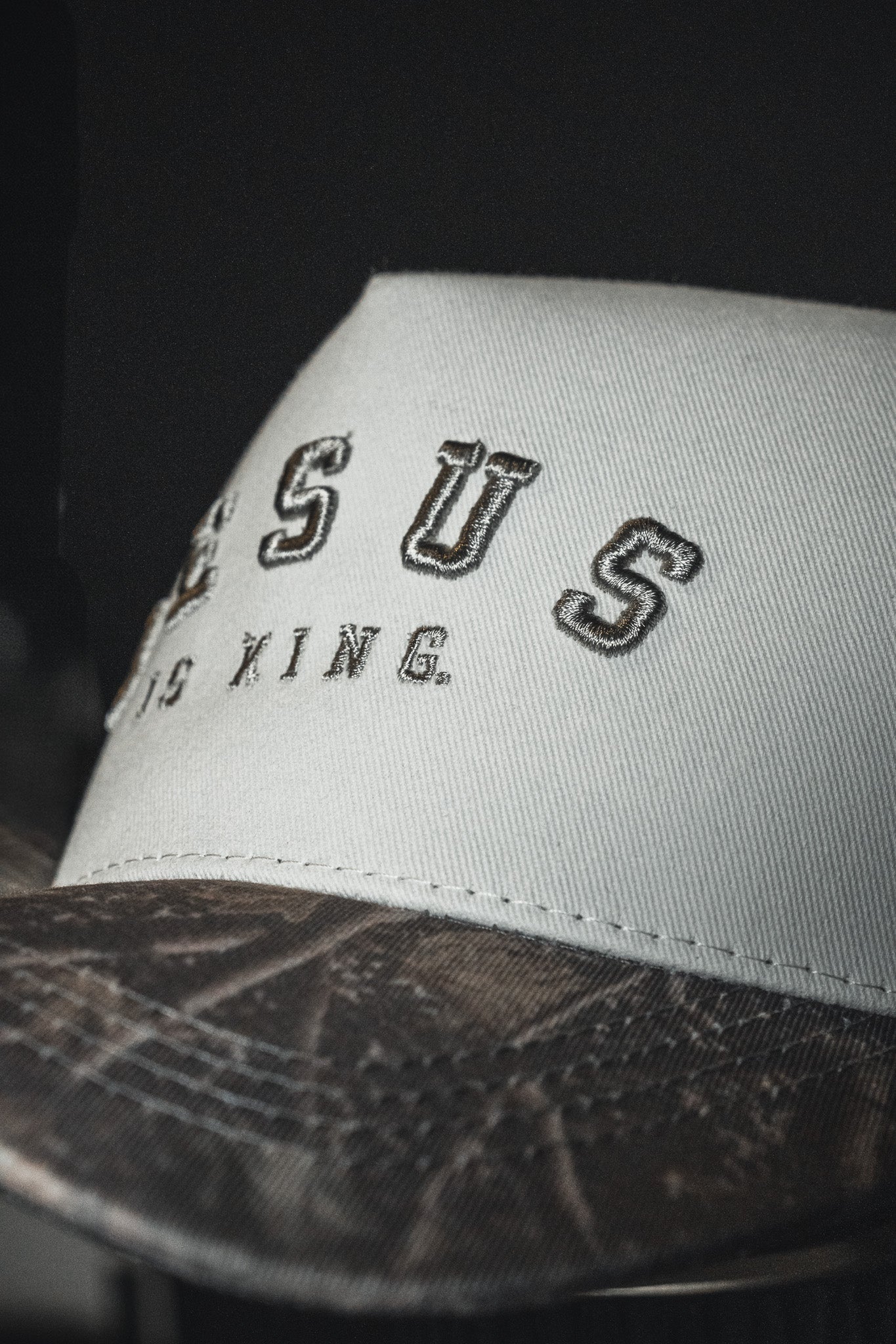 A close-up of the Born United Private Label Jesus is King - Ivory and Deadwood Camo Snapback, featuring JESUS IS KING boldly embroidered on the white crown and a patterned brown brim inspired by Deadwood Camo, set against a dark background.