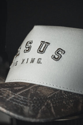 A close-up of the Born United Private Label Jesus is King - Ivory and Deadwood Camo Snapback, featuring JESUS IS KING boldly embroidered on the white crown and a patterned brown brim inspired by Deadwood Camo, set against a dark background.