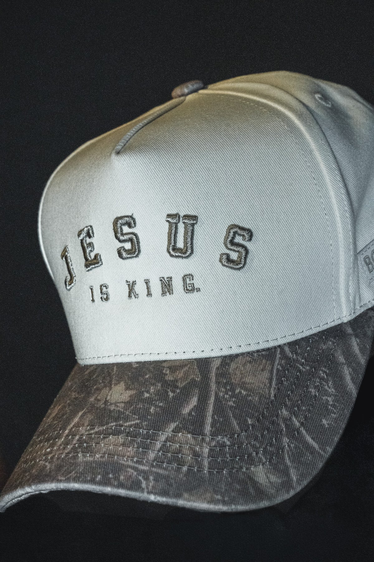 The Born United Private Label Jesus is King - Ivory and Deadwood Camo Snapback features “JESUS IS KING.” embroidered on the front, an ivory crown, and a Deadwood Camo hat-style brim, set against a black background.