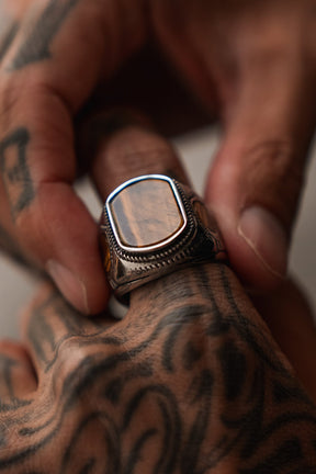 A close-up shows tattooed hands adjusting The Desert Relic Ring by Relentless Betrayal, highlighting its reflective rectangular stone set atop one finger.