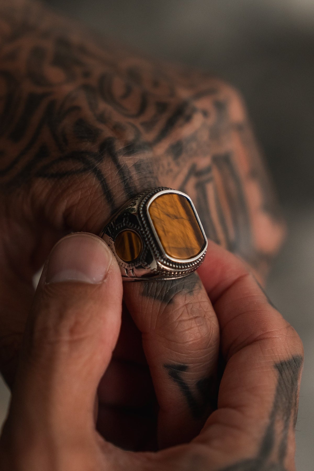 A tattooed hand holds The Desert Relic Ring by Relentless Betrayal—a silver, handcrafted ring showcasing a large rectangular amber or tiger’s eye stone, with another similar stainless steel ring visible on one finger.