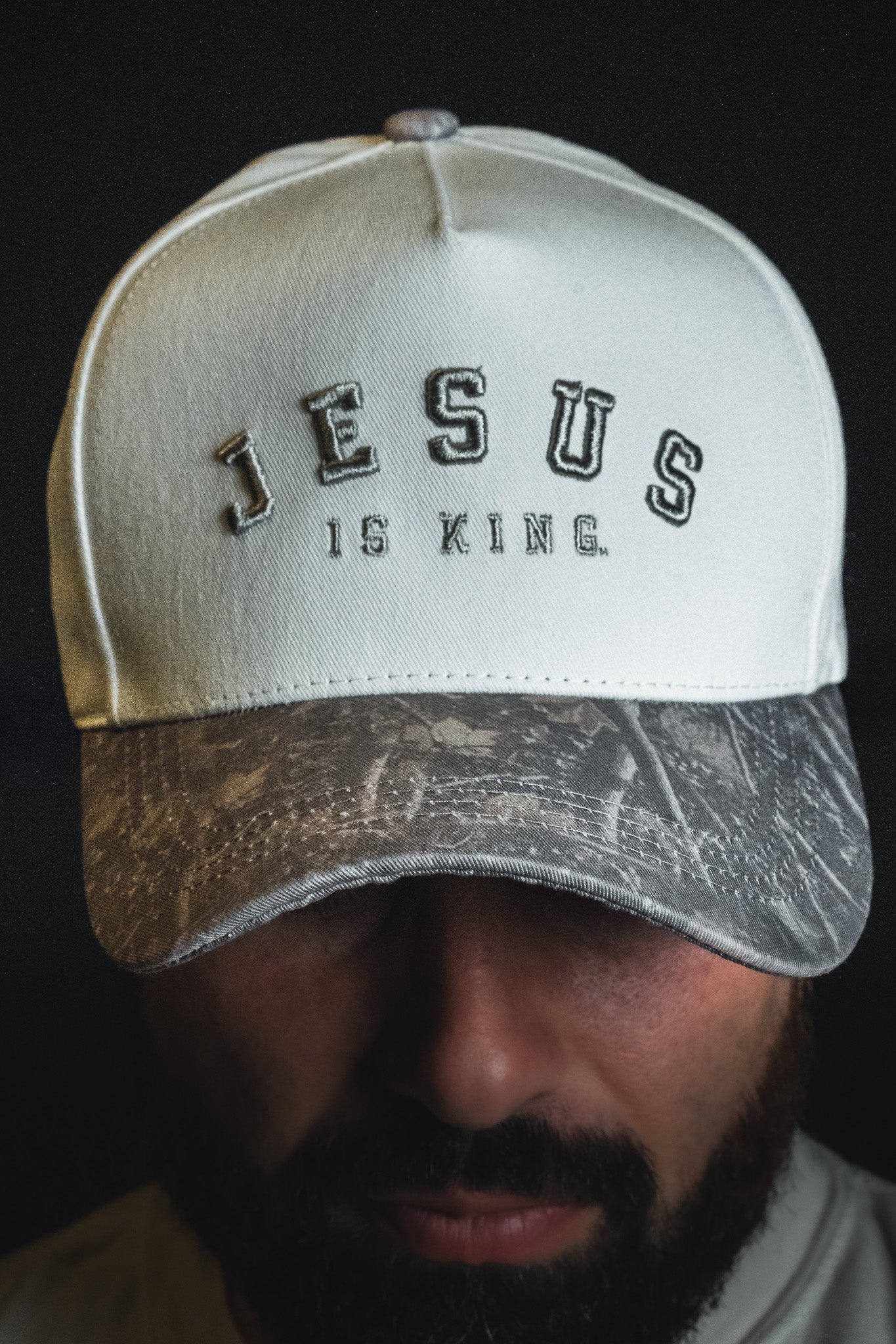 A man wearing a Born United Private Label “Jesus is King - Ivory and Deadwood Camo Snapback,” the cap partially shading his face.