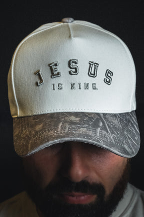 A man wearing a Born United Private Label “Jesus is King - Ivory and Deadwood Camo Snapback,” the cap partially shading his face.