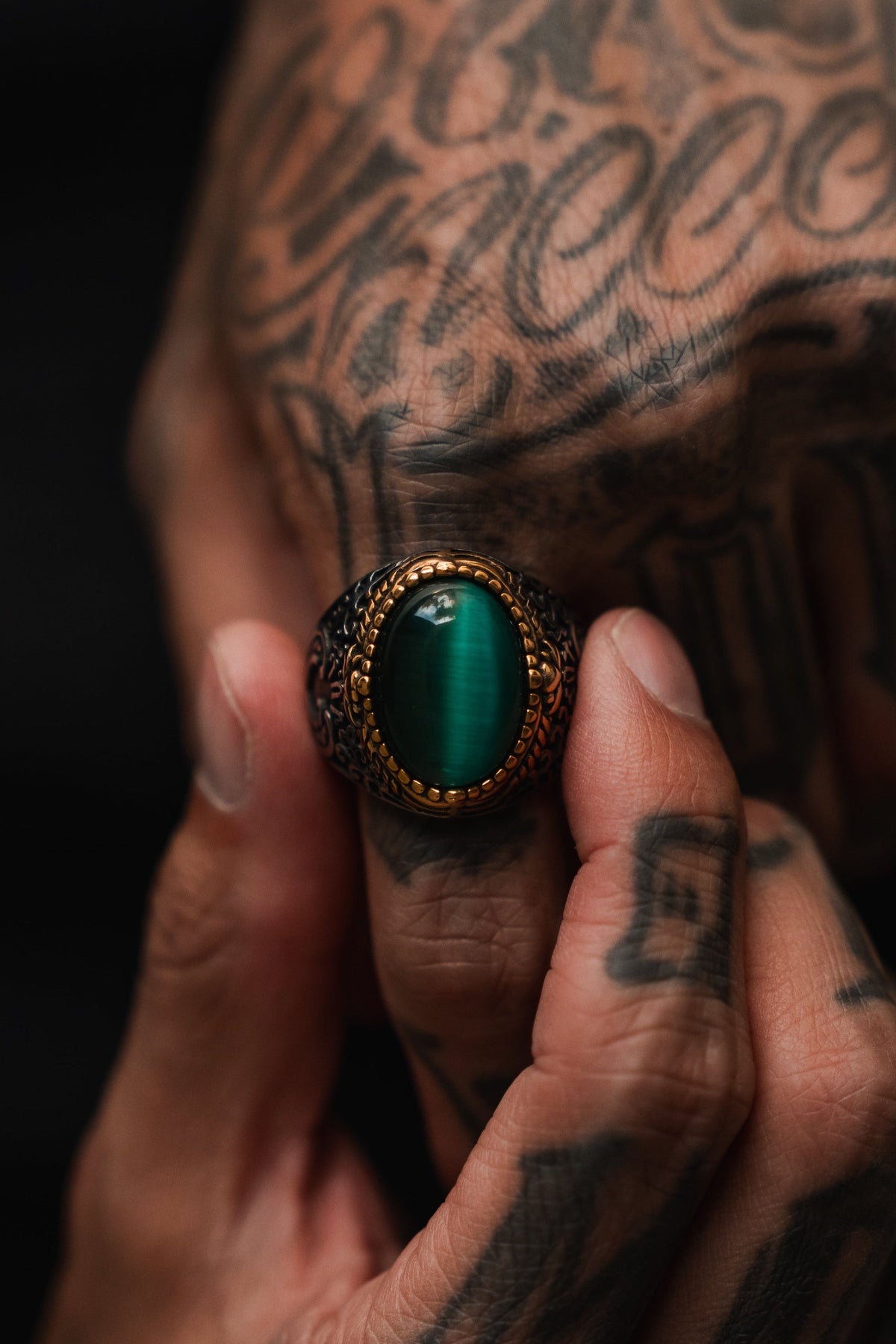 A tattooed hand holds The Relic Signet Ring by Relentless Betrayal, featuring an ornate gold setting and a large green oval stone at its center; its weathered finish stands out against the dark background.