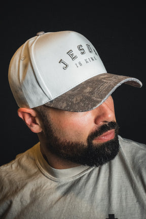 A bearded man in a light-colored shirt stands out against a black background, wearing the Born United Private Label “Jesus is King - Ivory and Deadwood Camo Snapback”—an embroidered baseball cap.