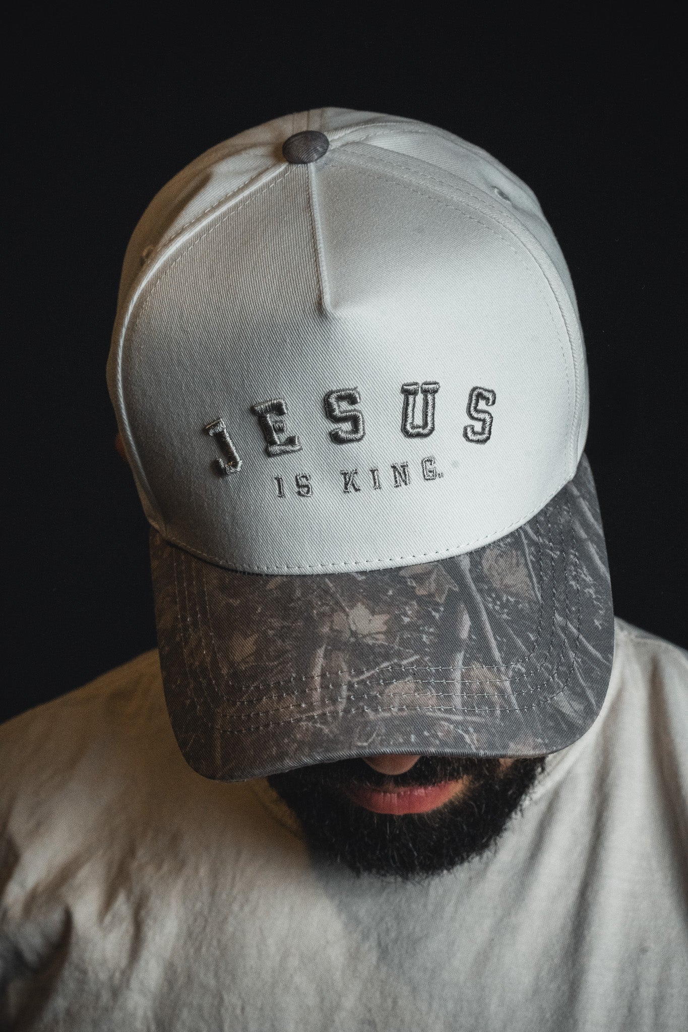 A person wears the Born United Private Label “Jesus is King - Ivory and Deadwood Camo Snapback” with their head tilted downward against a dark background.
