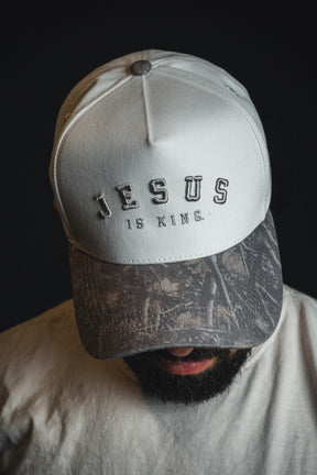 A person wears the Born United Private Label “Jesus is King - Ivory and Deadwood Camo Snapback” with their head tilted downward against a dark background.