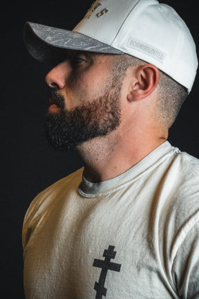 A man with a trimmed beard is shown in profile against a dark background, wearing the Born United Private Label “Jesus is King - Ivory and Deadwood Camo Snapback” and a light-colored shirt featuring a black cross symbol.