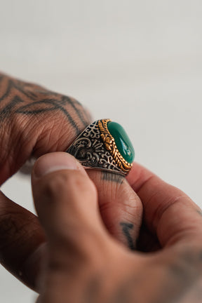A tattooed hand wears The Relic Signet Ring by Relentless Betrayal, showcasing its ornate designs, large deep stone center, and weathered silver finish as another hand adjusts the ring.