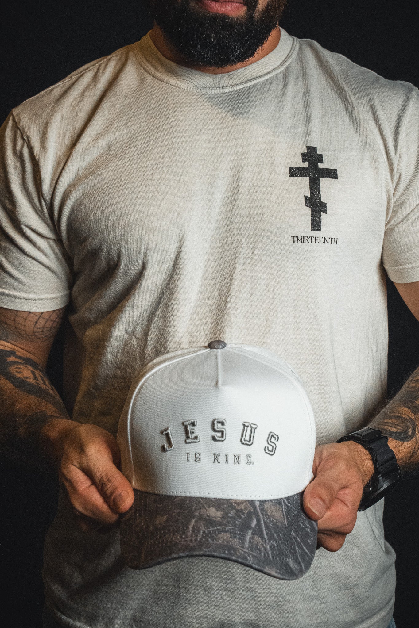 A man in a white t-shirt with a cross graphic holds the Jesus is King - Ivory and Deadwood Camo Snapback by Born United Private Label, featuring bold white and gray text embroidered on the front.