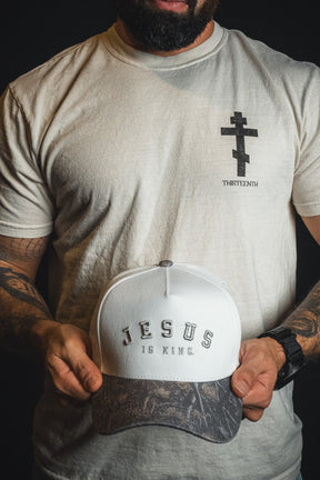 A man in a white t-shirt with a cross graphic holds the Jesus is King - Ivory and Deadwood Camo Snapback by Born United Private Label, featuring bold white and gray text embroidered on the front.