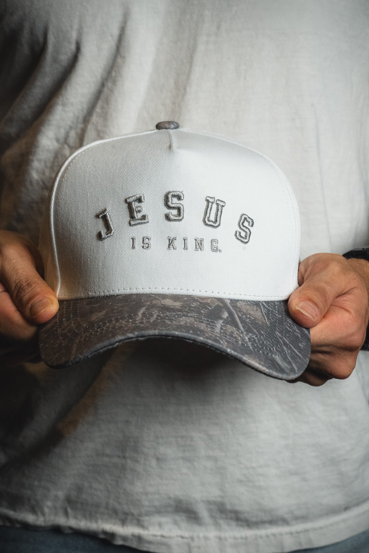 A person in a white shirt holds the Born United Private Label “Jesus is King - Ivory and Deadwood Camo Snapback,” featuring the words JESUS IS KING. embroidered on the front.