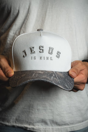 A person in a white shirt holds the Born United Private Label “Jesus is King - Ivory and Deadwood Camo Snapback,” featuring the words JESUS IS KING. embroidered on the front.