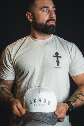 A bearded man wearing a light shirt with a cross symbol holds the Born United Private Label “Jesus is King - Ivory and Deadwood Camo Snapback” against a dark background.