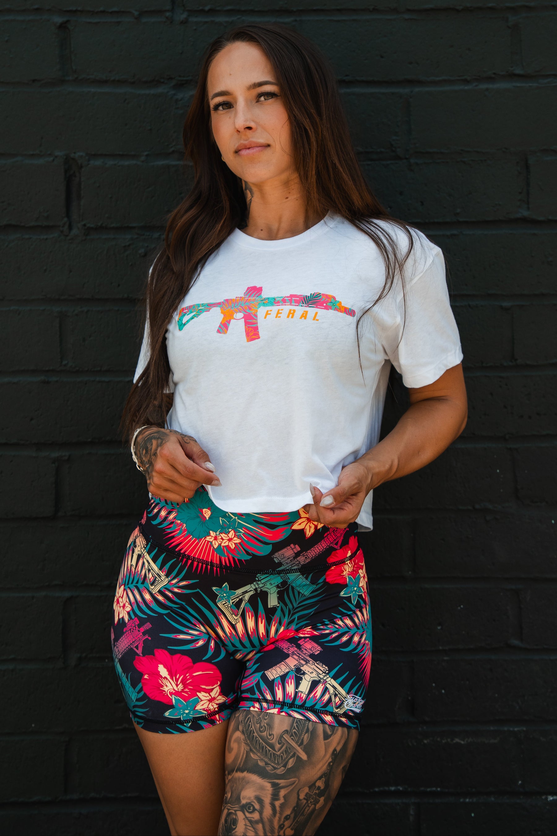 A woman with long hair and a leg tattoo stands by a dark brick wall, wearing the Tango Charlie Apparel Feral Women's White Crop—a t-shirt with a colorful graphic and "FERAL" text—and high-waisted floral shorts.