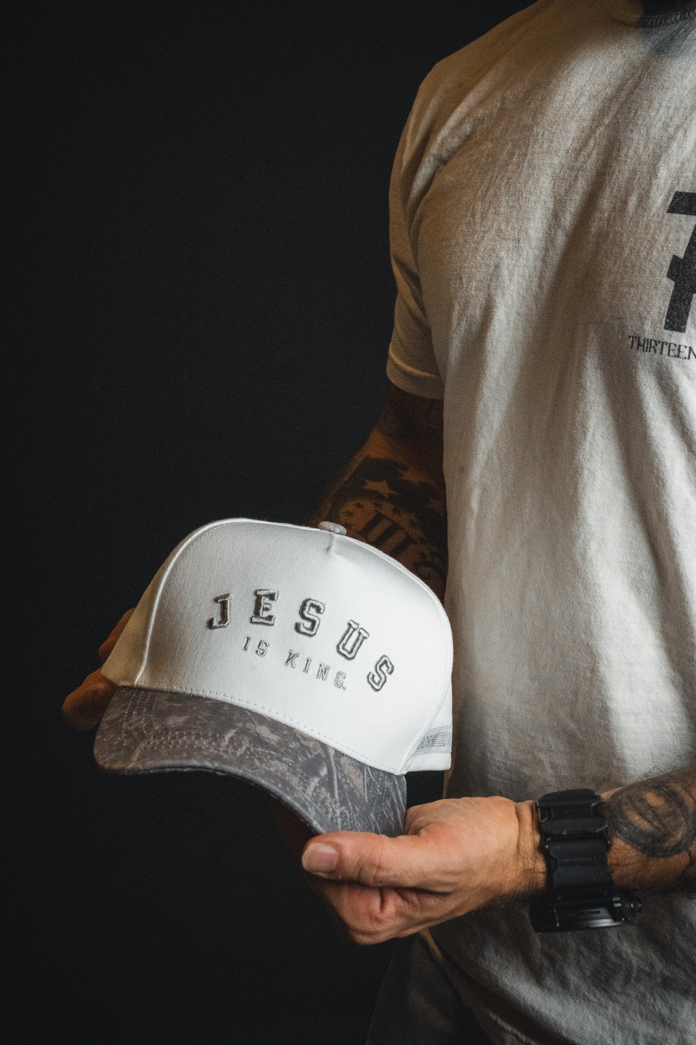A person wearing a white t-shirt and black watch holds the Born United Private Label “Jesus is King - Ivory and Deadwood Camo Snapback” embroidered baseball cap.