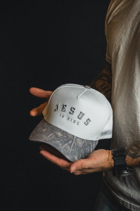 A person wearing a white shirt and black watch holds the Born United Private Label “Jesus is King - Ivory and Deadwood Camo Snapback.”.