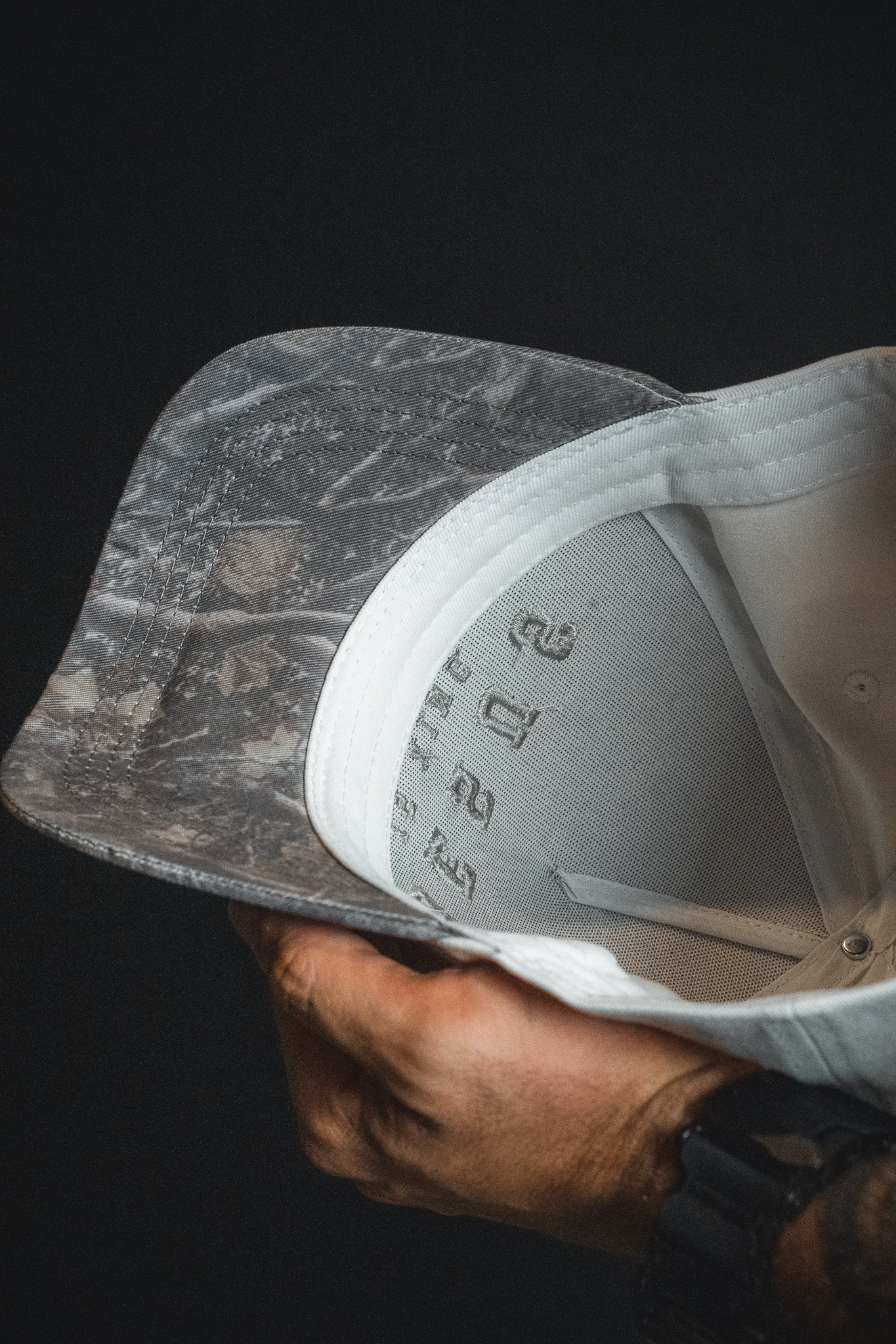 A hand holding a Jesus is King - Ivory and Deadwood Camo Snapback from Born United Private Label, showing the inside lining and embroidered numbers under the brim of this unique baseball cap.