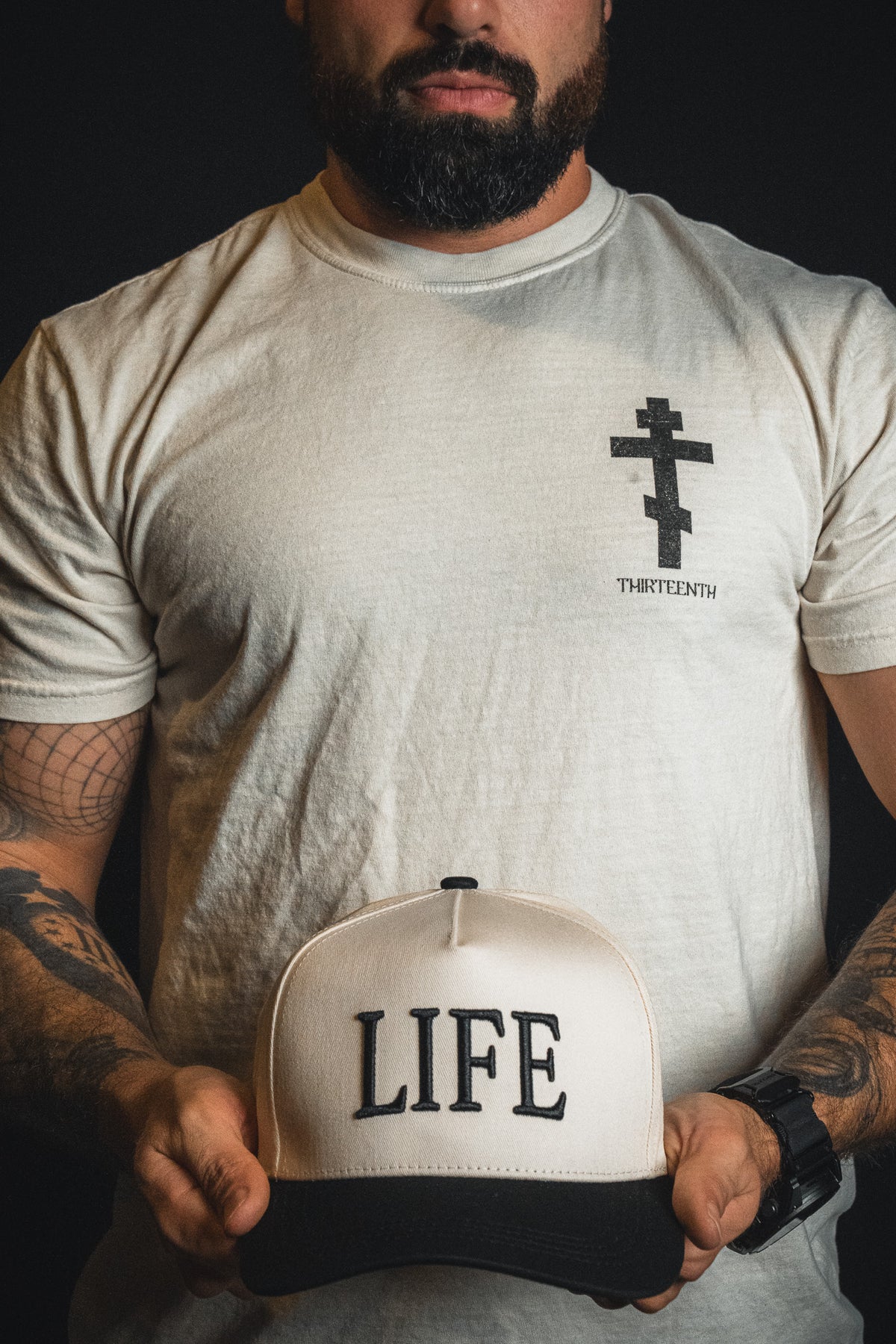 A man with tattoos and a beard holds the LIFE - Ivory and Black Snapback from Born United Private Label while wearing a shirt featuring a cross symbol and “THIRTEENTH” printed on it.