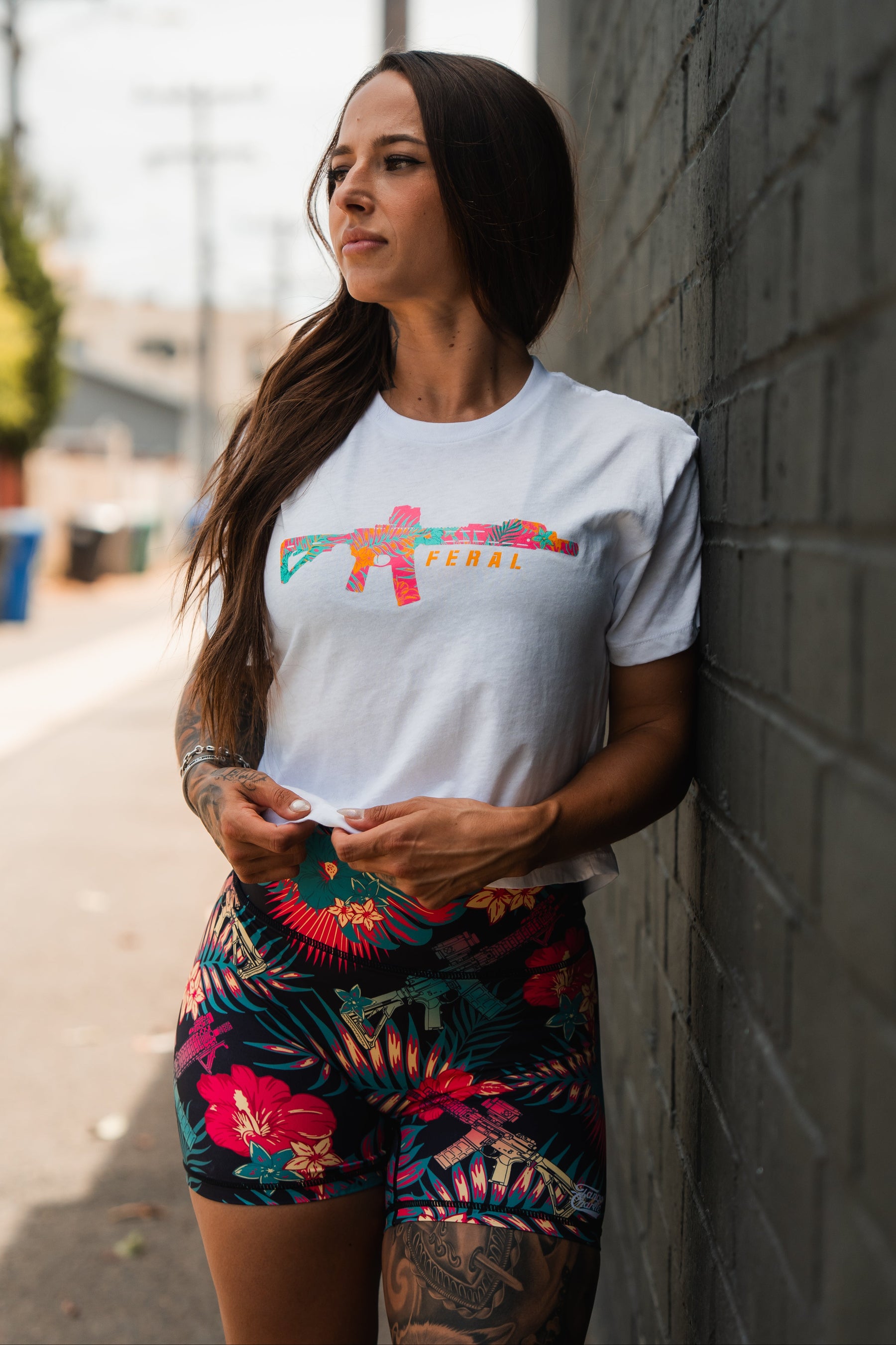 A woman with long brown hair stands against a dark brick wall, wearing the Tango Charlie Apparel "Feral" Women's White Crop, paired with patterned tropical shorts. She looks to the side, holding her shirt hem.