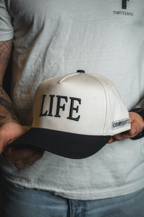 A person holding the LIFE - Ivory and Black Snapback by Born United Private Label, featuring LIFE embroidered on the front and BORN UNITED on the side—a stylish hat with a spiritual message perfect for those seeking inspiration.