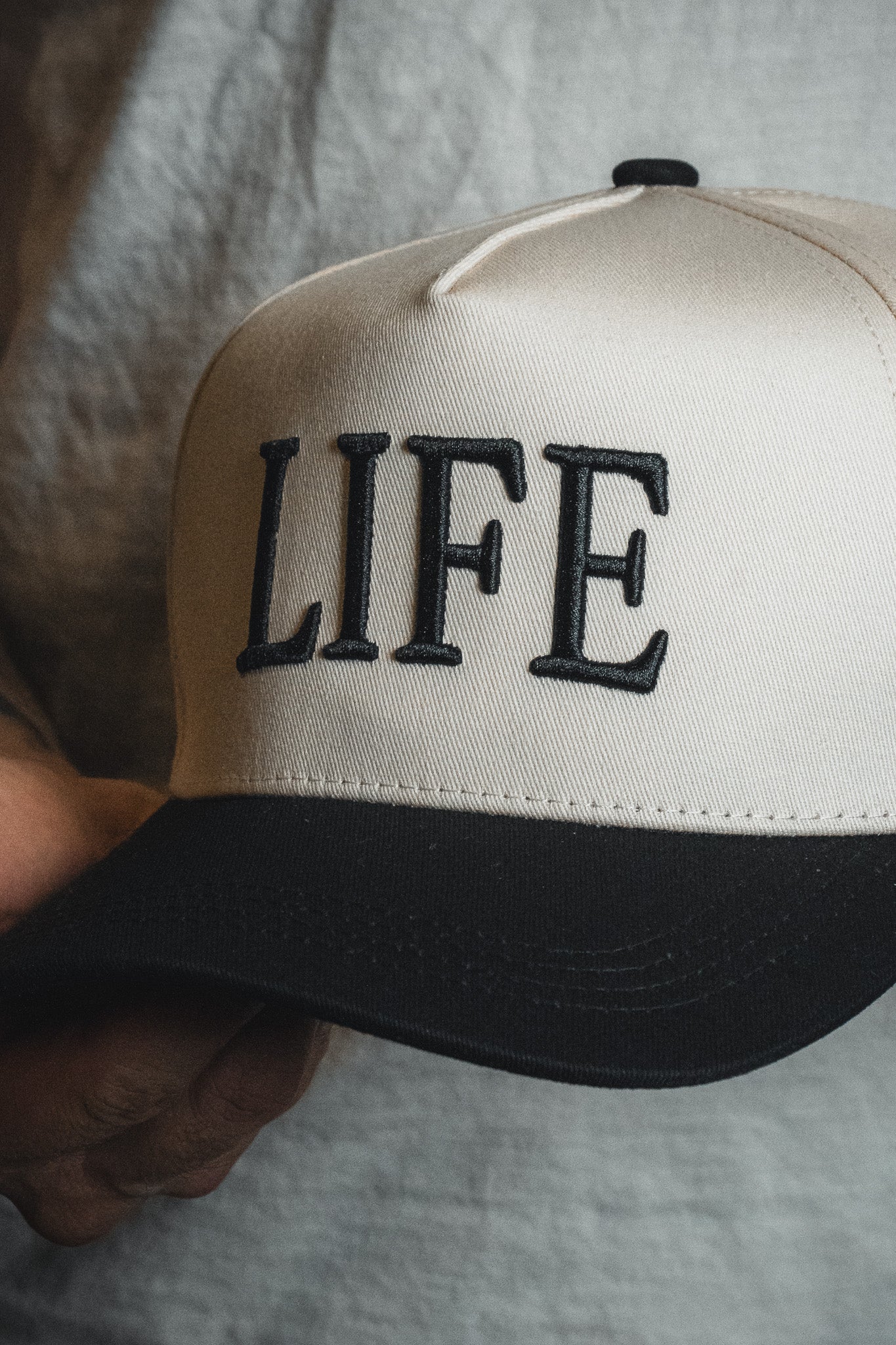 A person holds the LIFE - Ivory and Black Snapback by Born United Private Label, featuring bold black embroidery of the word “LIFE” on the front, making this cap a striking spiritual statement.