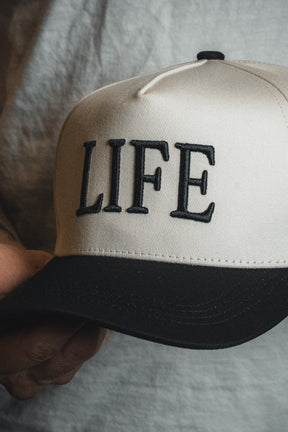 A person holds the LIFE - Ivory and Black Snapback by Born United Private Label, featuring bold black embroidery of the word “LIFE” on the front, making this cap a striking spiritual statement.