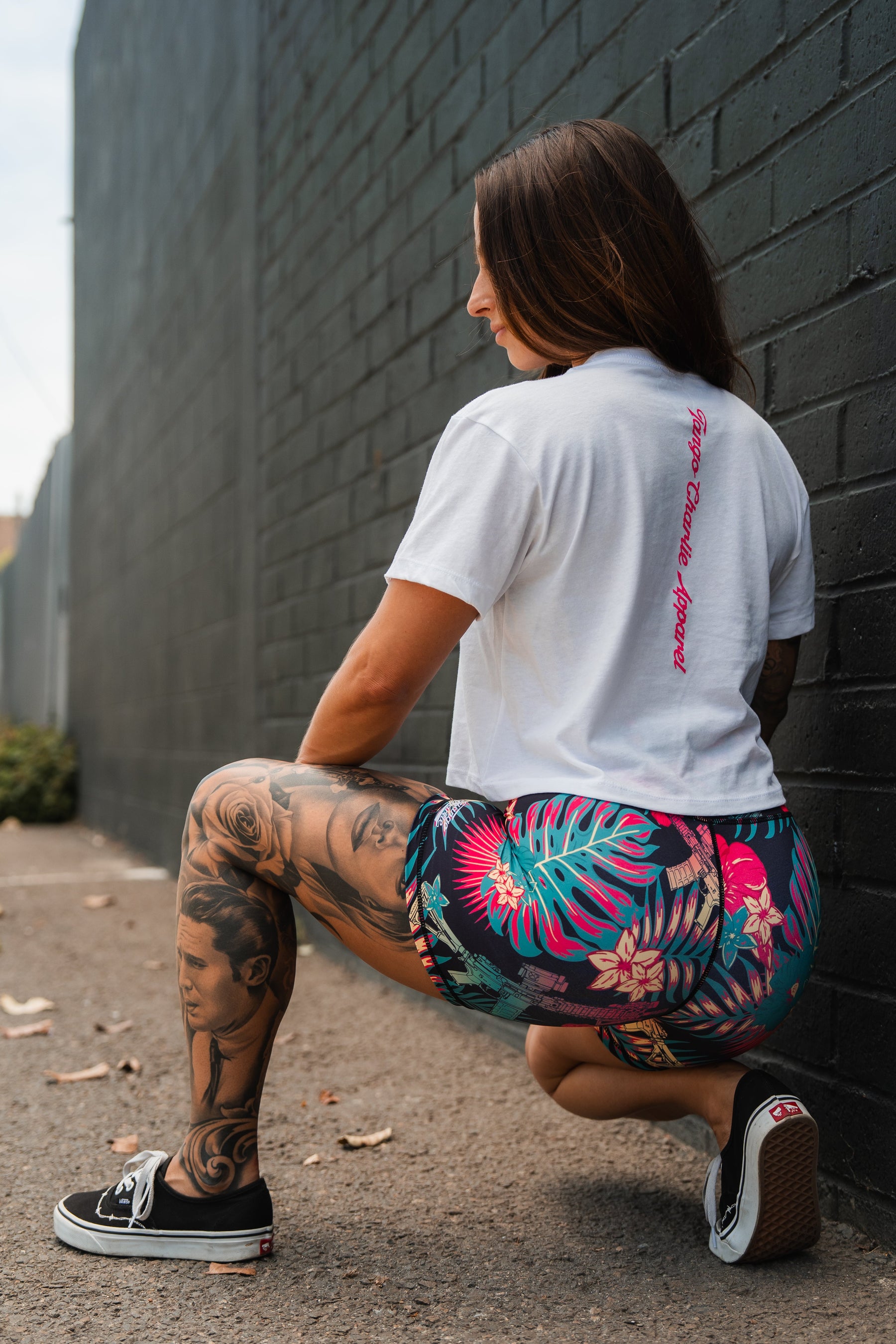 Against a dark brick wall, a woman with long brown hair and leg tattoos squats outdoors wearing the Feral - Women's White Crop by Tango Charlie Apparel, paired with floral shorts and black Vans.