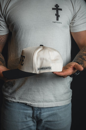 A person with tattooed arms holds the LIFE - Ivory and Black Snapback from Born United Private Label, wearing a white t-shirt featuring a cross and the word “THIRTEENTH.”.