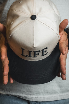 A person holds the Born United Private Label LIFE - Ivory and Black Snapback, featuring bold “LIFE” lettering on the front for a stylish spiritual statement.