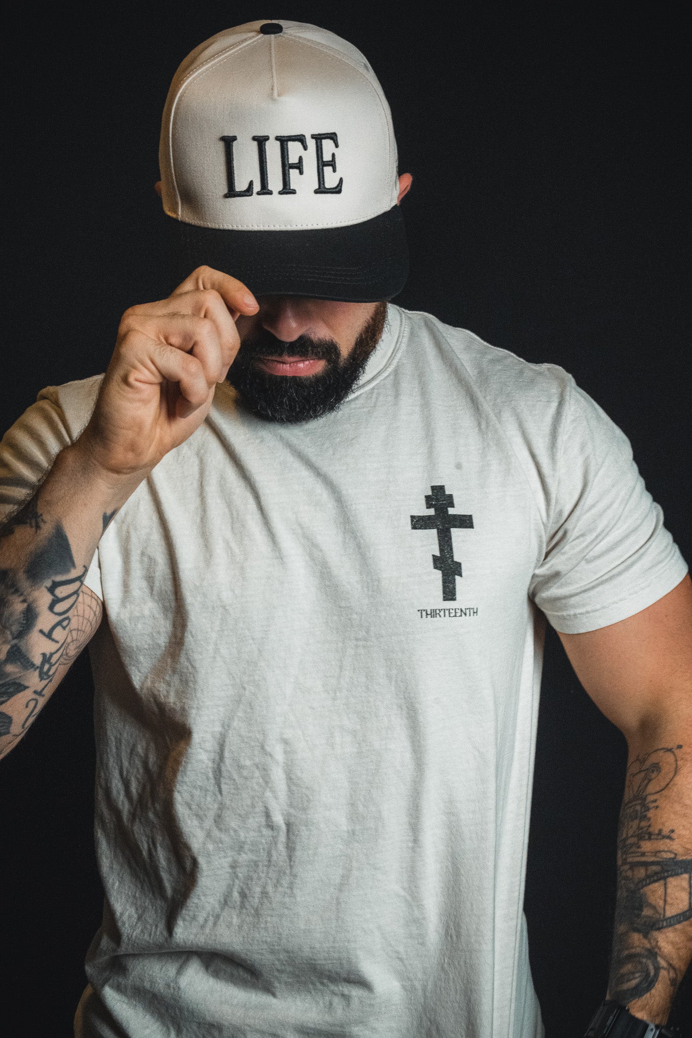 A tattooed man adjusts a Born United Private Label LIFE - Ivory and Black Snapback—a snapback hat featuring a spiritual message—while wearing a white shirt with a cross design on the left side.