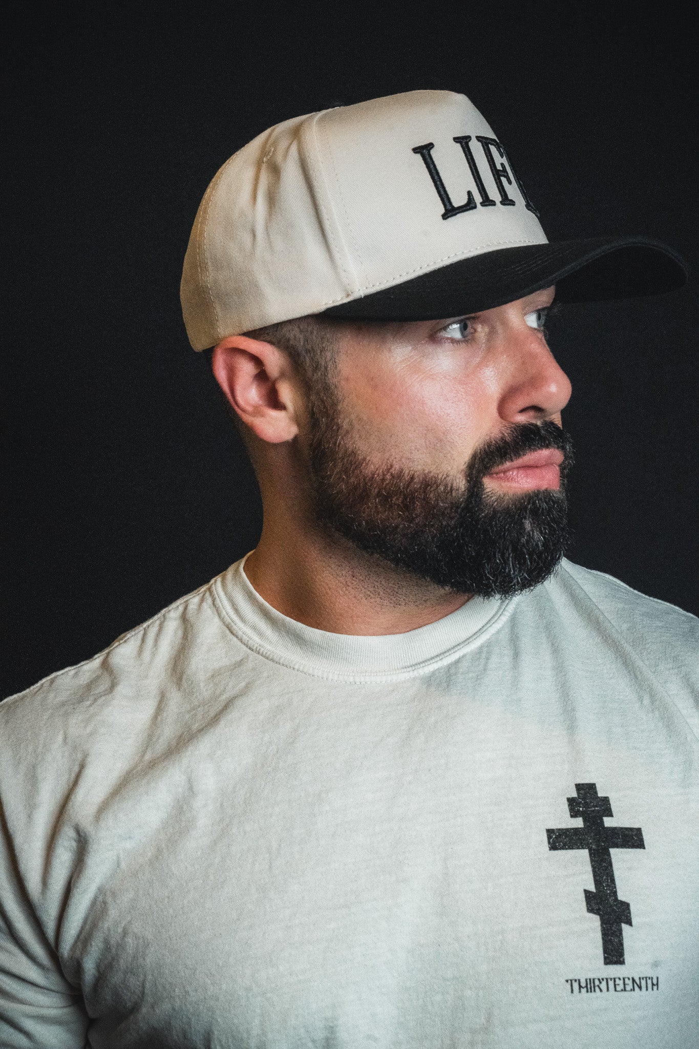 A bearded man, wearing the Born United Private Label “LIFE - Ivory and Black Snapback” and a white shirt with a black cross design, looks to the side against a dark background.