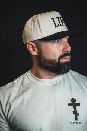 A bearded man, wearing the Born United Private Label “LIFE - Ivory and Black Snapback” and a white shirt with a black cross design, looks to the side against a dark background.