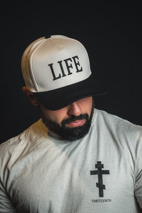 A bearded man wearing the Born United Private Label “LIFE - Ivory and Black Snapback” and a T-shirt with a cross and the word THIRTEENTH looks down thoughtfully against a dark background.
