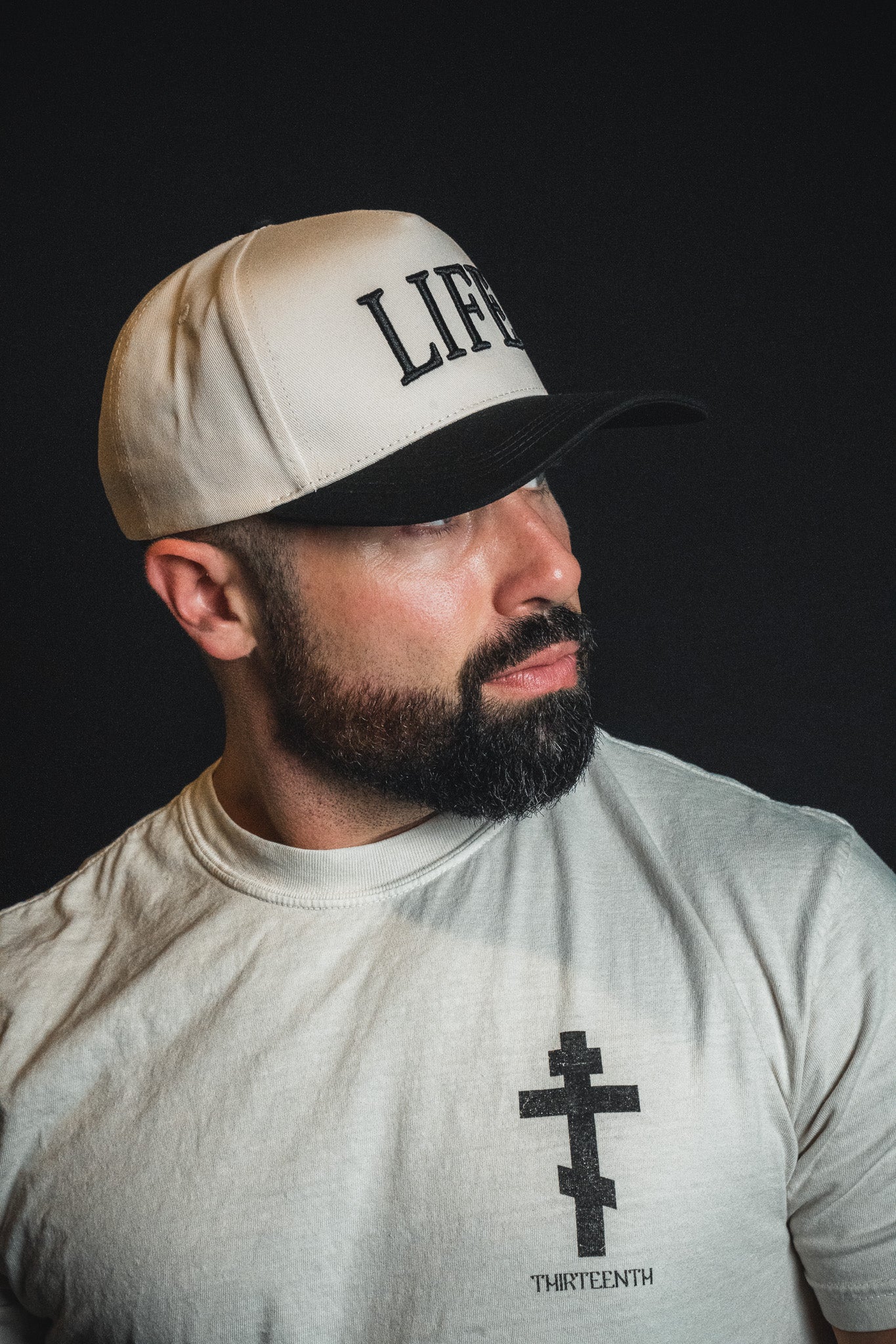 A man with a beard, looking to the side against a dark background, wears the Born United Private Label “LIFE - Ivory and Black Snapback” and a white t-shirt featuring a cross design.
