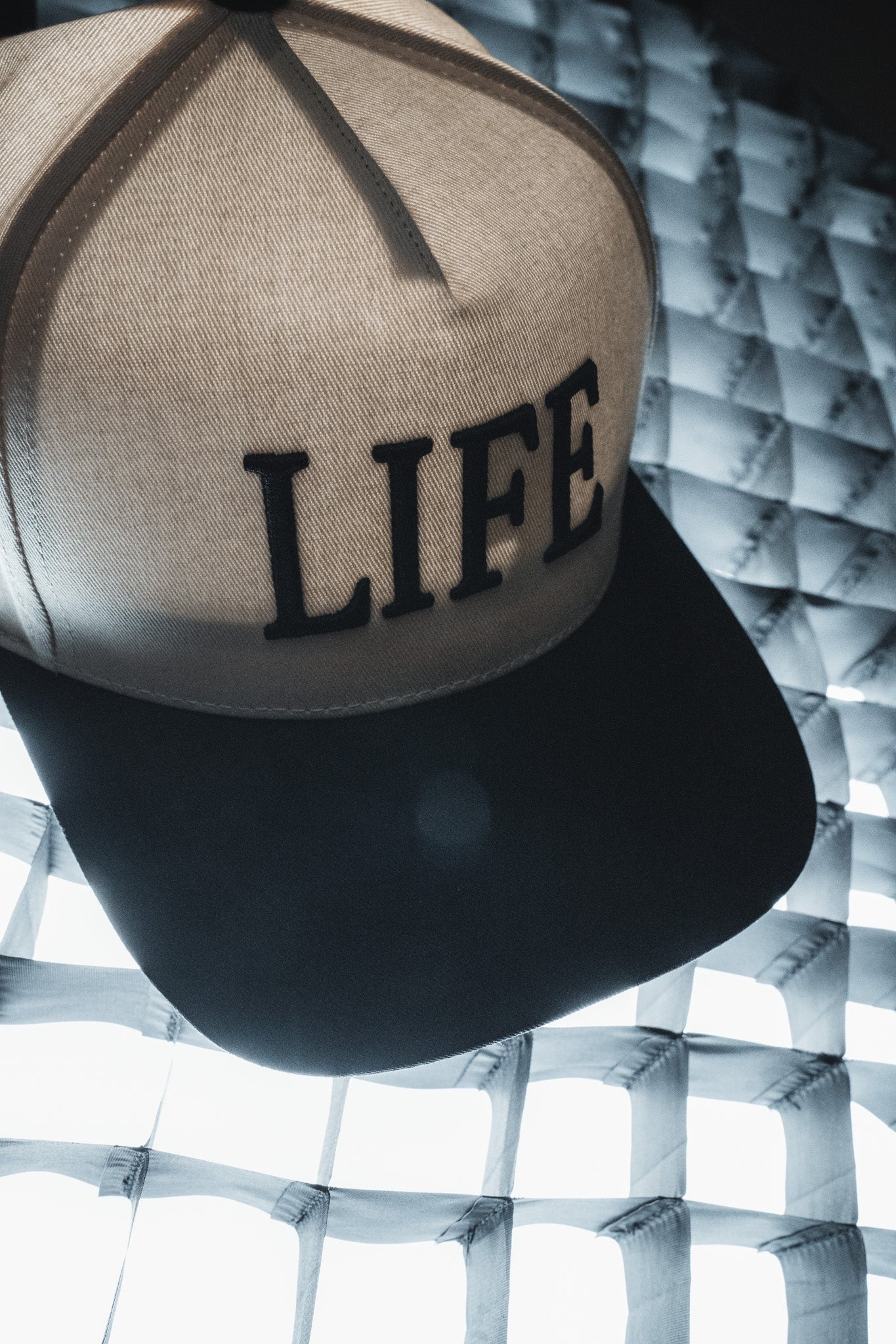 A Born United Private Label LIFE - Ivory and Black Snapback featuring embroidered lettering on the front sits atop a grid-patterned, glowing surface, offering a stylish hat with a subtle spiritual message for those who value both meaning and design.