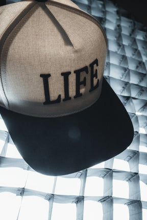 A Born United Private Label LIFE - Ivory and Black Snapback featuring embroidered lettering on the front sits atop a grid-patterned, glowing surface, offering a stylish hat with a subtle spiritual message for those who value both meaning and design.